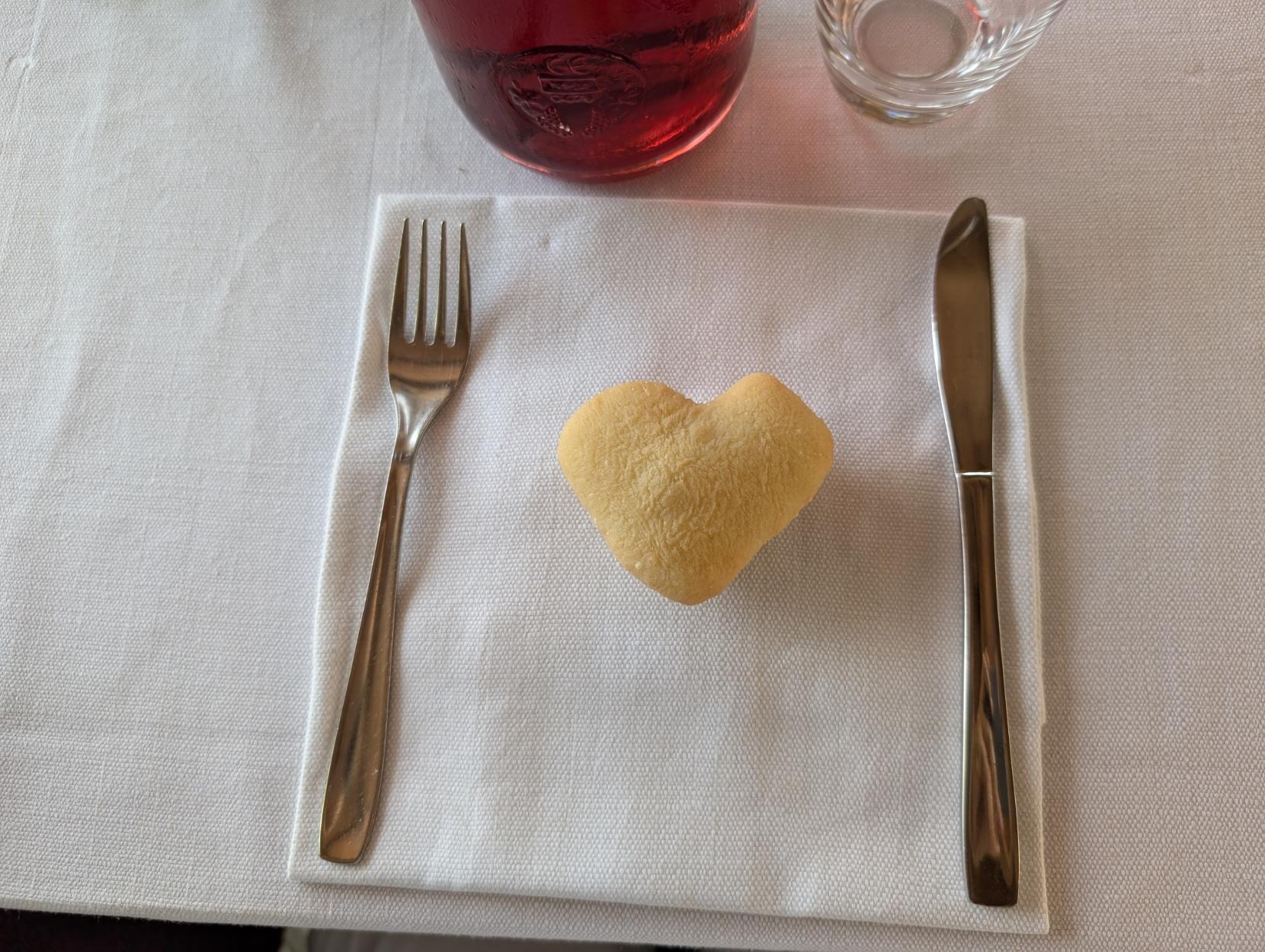 A top-down shot shows a heart-shaped piece of bread resting on a white cloth napkin, positioned between a silver fork on the left and a silver knife on the right. In the background, a bottle filled with a red wine is visible, along with an empty clear glass.