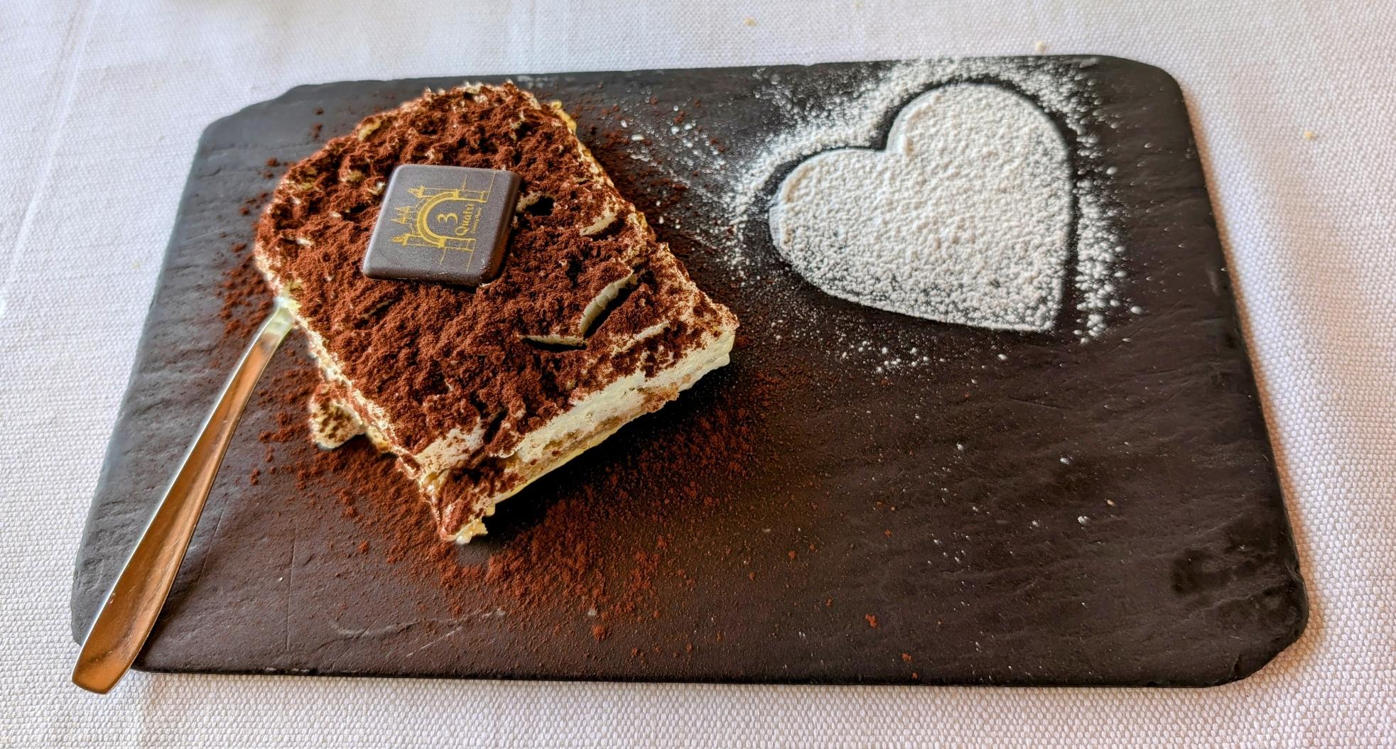 A high-angle shot captures a slice of tiramisu presented on a dark rectangular slate. The tiramisu is heavily dusted with cocoa powder and topped with a small, square piece of chocolate. To the right of the dessert, a heart shape is dusted onto the slate with powdered sugar, and a small spoon rests beside the tiramisu.
