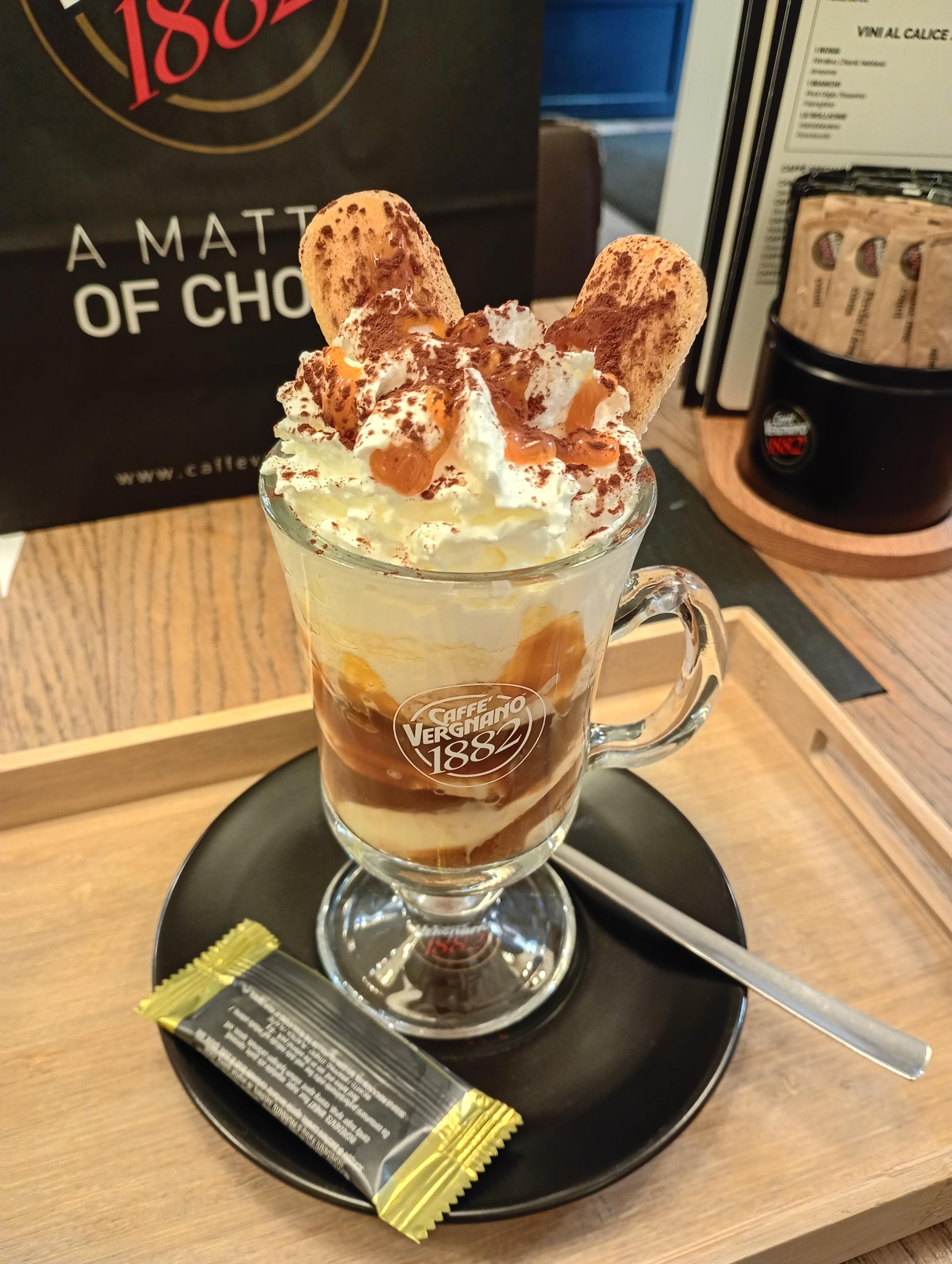 A photo of a tall glass with the coffee based drink in the style of Tiramisu - with 2 ladyfingers poking out of the whipped cream.