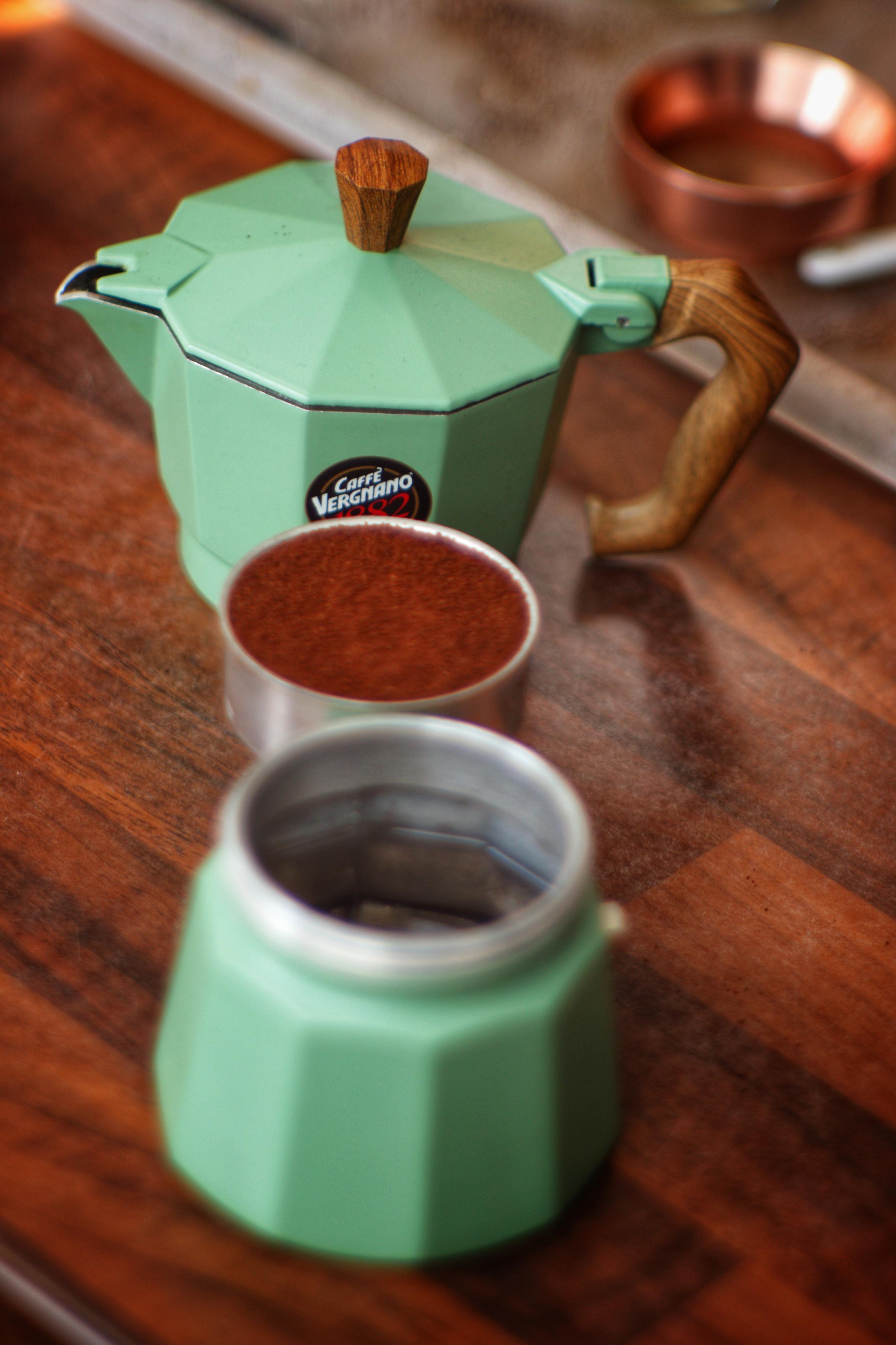 A mint-green stovetop moka pot with a wooden handle and knob sits on a wooden table, taken apart beside a metal filter basket filled with ground coffee, with a blurred copper coffee alignment tool in the background.