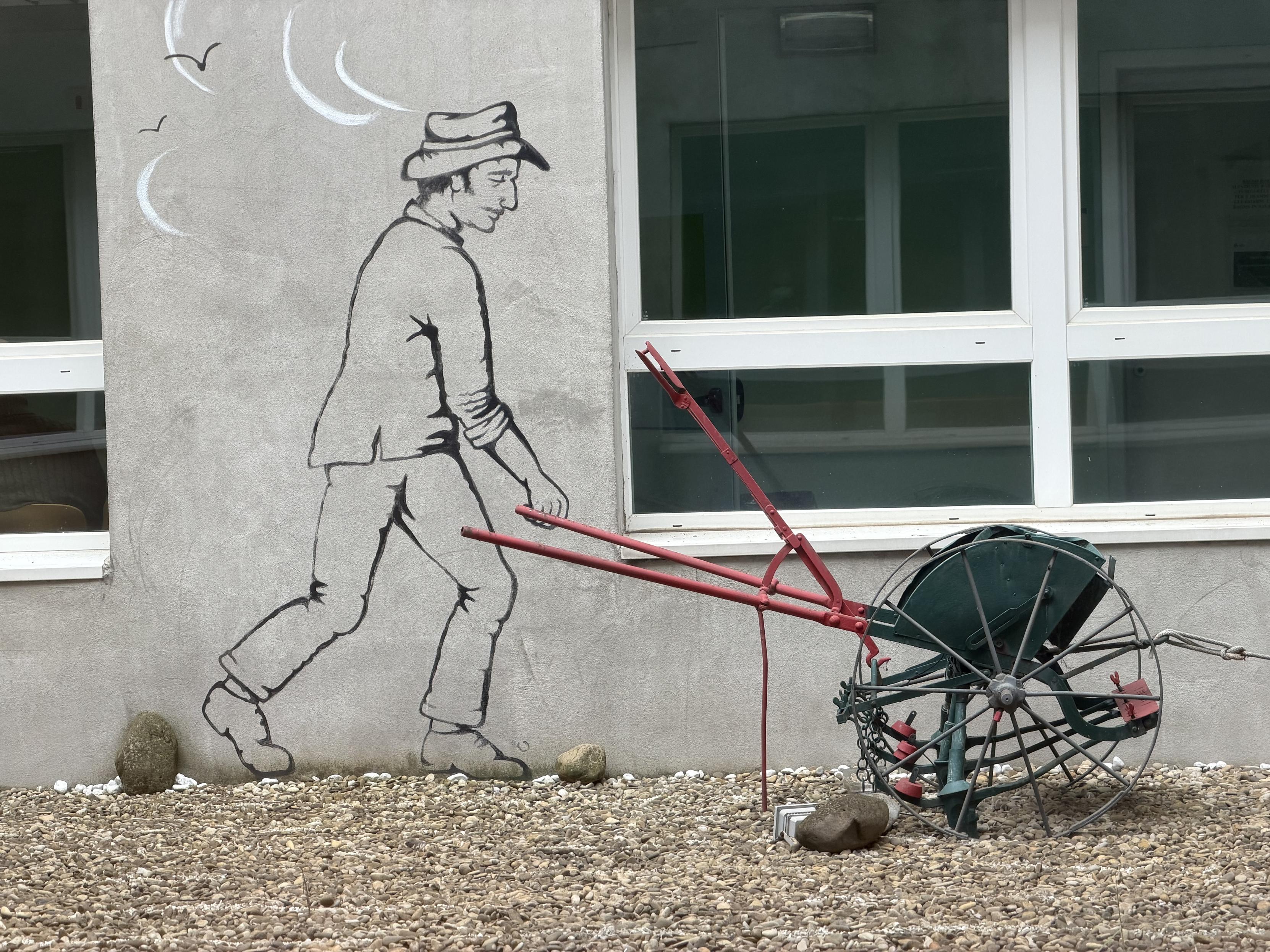 A black line mural of a farmer in a hat walking and pushing a plow is painted on a gray wall, aligned so that the drawing appears to push a real vintage red and green plow placed in front of the building. The ground is covered with small stones, and a window reflects light behind the scene.