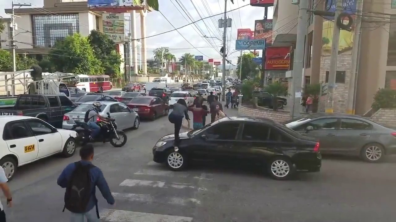 A car has stopped well beyond the middle of a crosswalk, so a pedestrian has chosen to walk over the hood of the car, disregarding any potential damage to the car caused by an adult walking on it