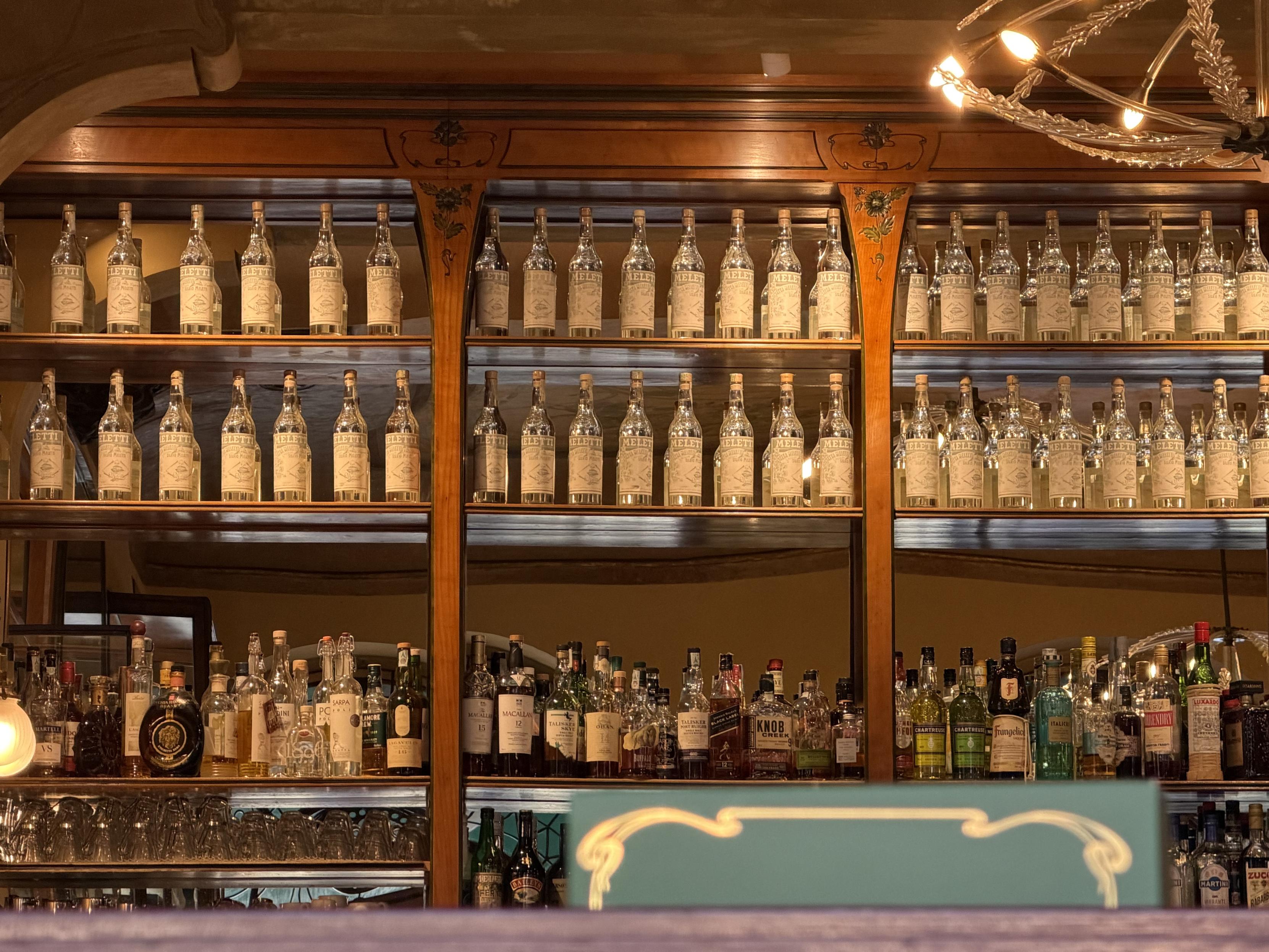Ornate wooden bar shelves lined with rows of empty Anisetta Meletti bottles above a large assortment of spirits, warmly lit by a decorative chandelier.