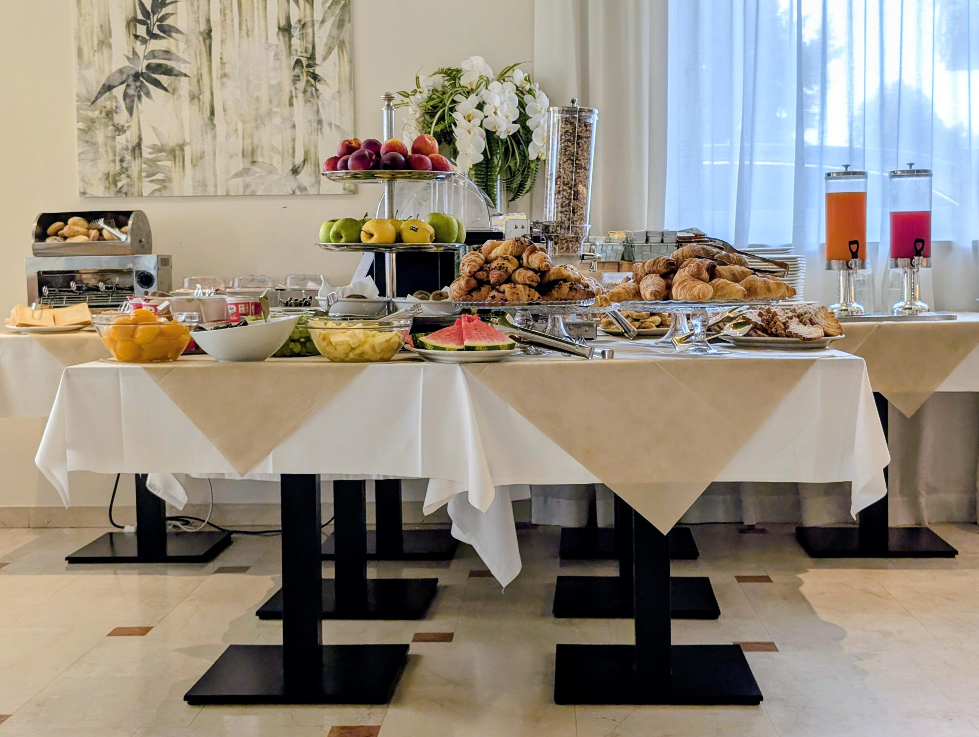 A full view of a buffet breakfast layout in a hotel dining area, with a white and beige table. Displayed are tiered levels of fruit, fresh croissants and baked goods, bowls with fresh grapes and melon, plus cereal in dispensers, and carafes of juice. A canvas painting and flowers are in the background.
