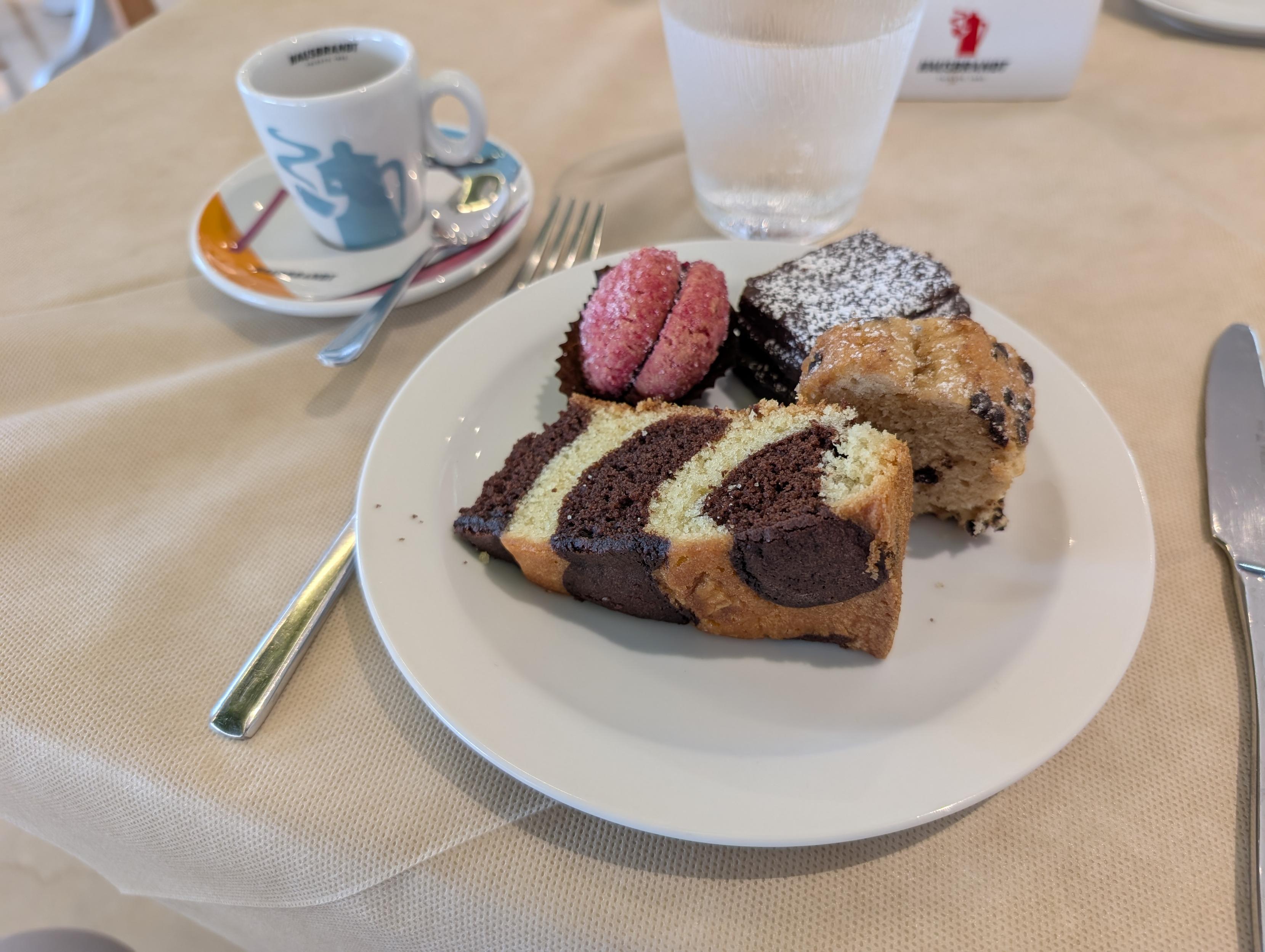 A close-up of a white plate with assorted breakfast pastries, including a slice of marble cake, a small chocolate cake sprinkled with powdered sugar, a pink cookie pastry, and a chocolate chip muffin. In the background, an espresso cup on a saucer, a glass of water, and cutlery sit on a beige tablecloth.