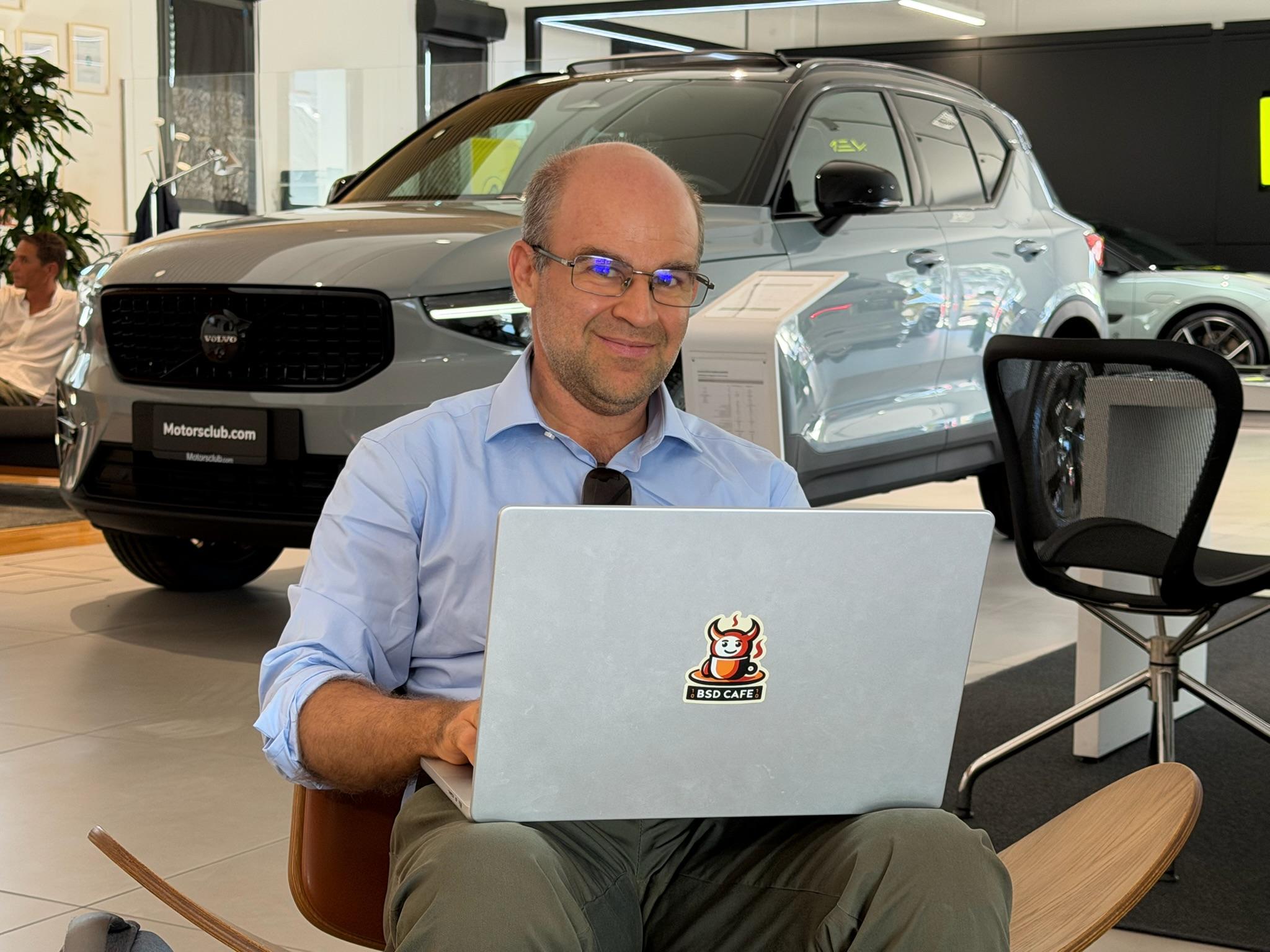 I'm sitting comfortably in a car showroom, smiling while working on a laptop with a BSD Cafe sticker on the lid. A grey Volvo is parked behind me, with other cars and showroom furniture visible in the background.