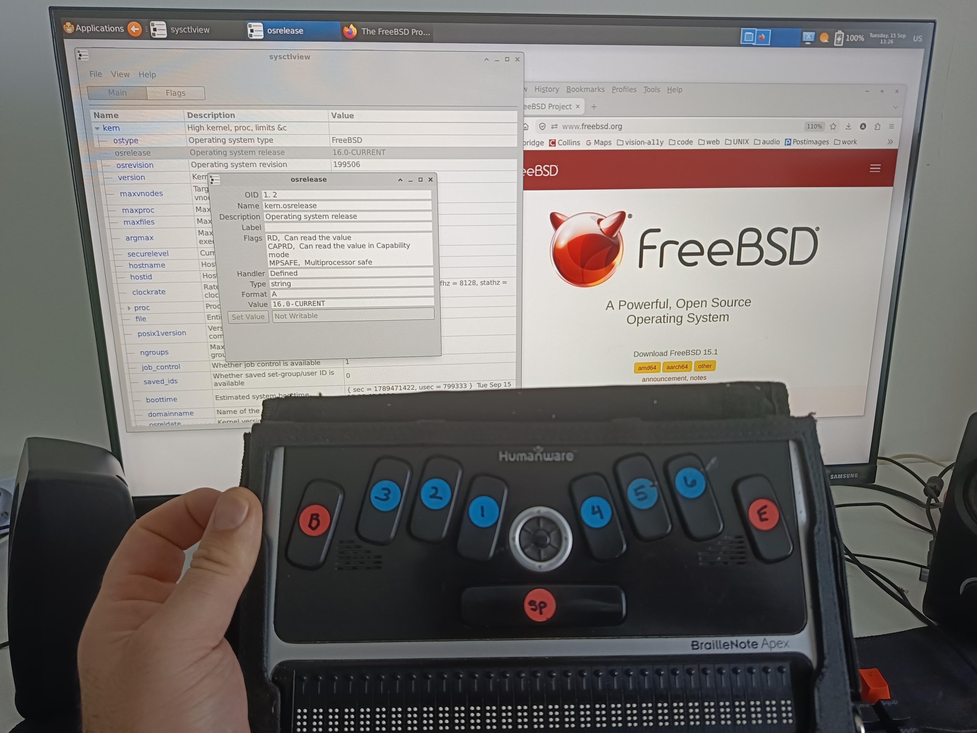 The HumanWare BrailleNote Apex photographed from above, with a monitor showing FreeBSD in the background.