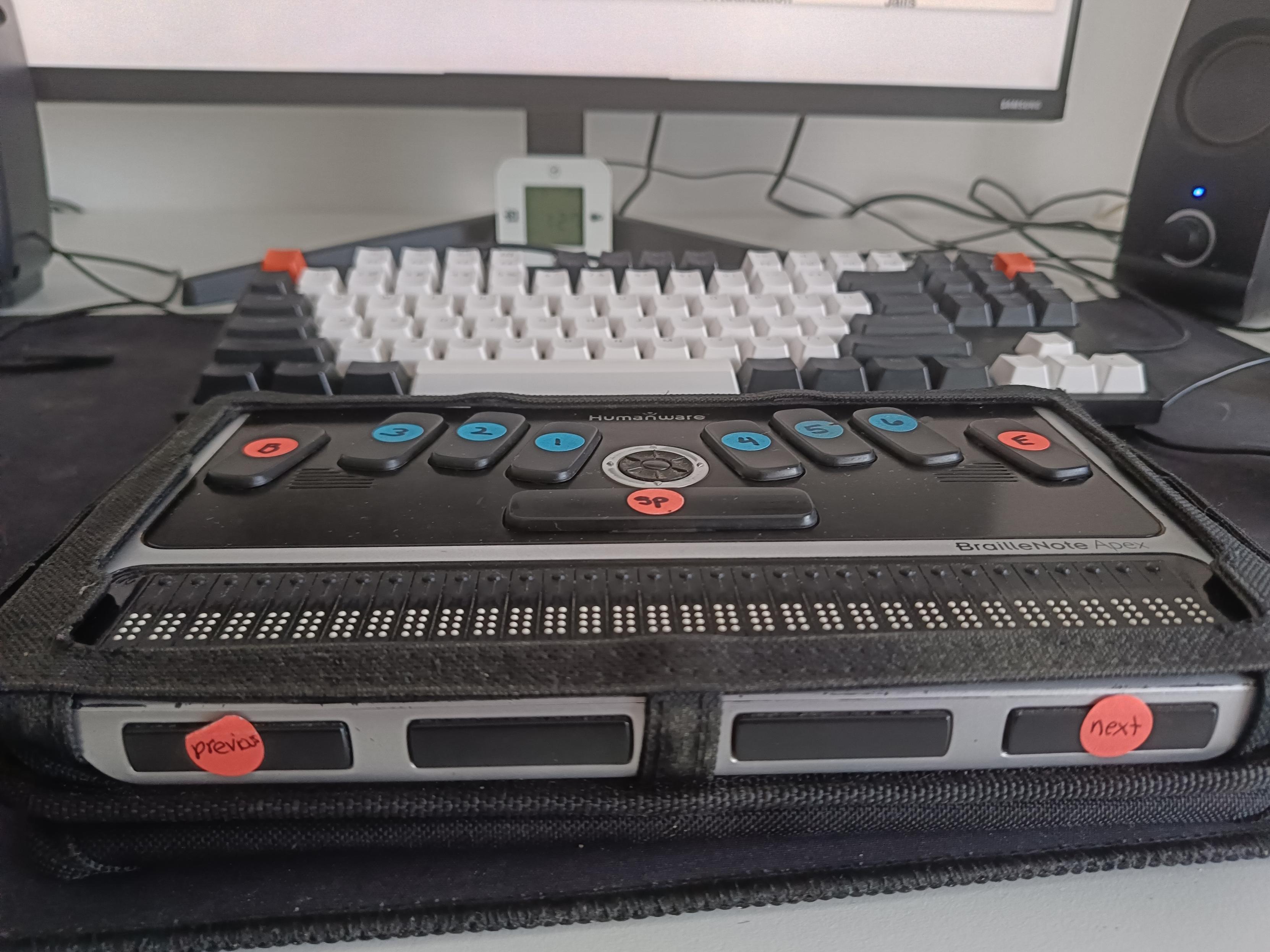 The HumanWare BrailleNote Apex photographed from the front, showing the Next and Previous buttons, with a monitor showing FreeBSD in the background.